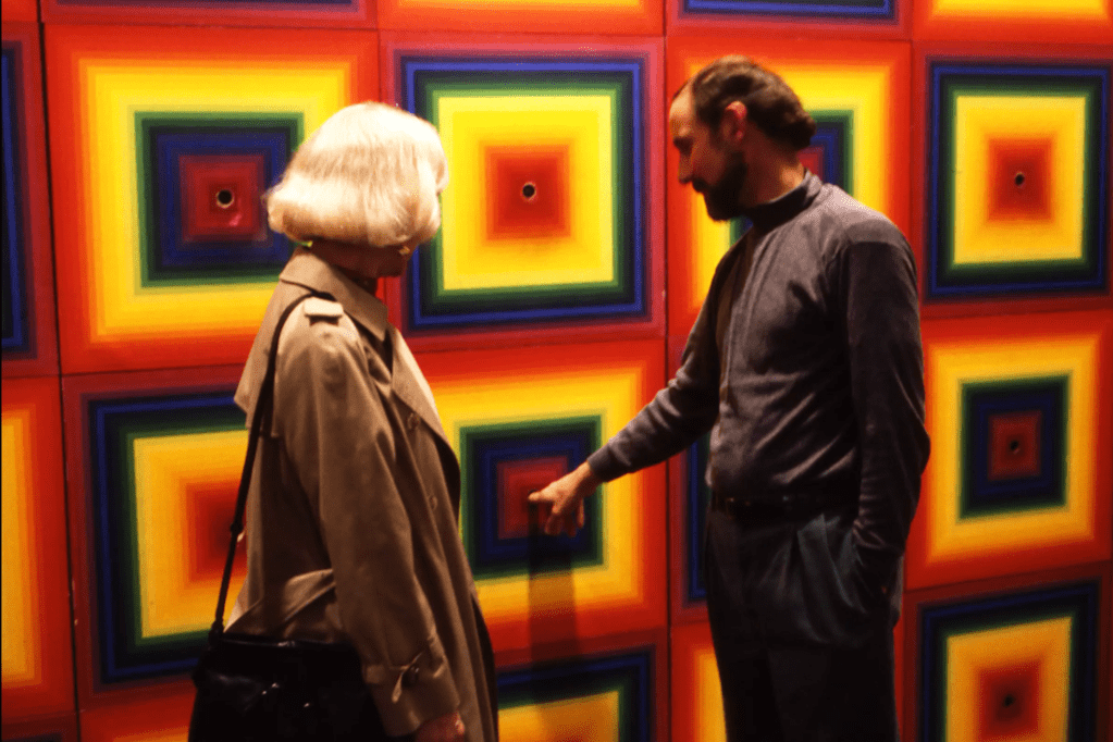 Two museum visitors interact with Ay-O's 'Rainbow Environment No. 7 ' installation. A person on the left with white blonde hair and a tan coat faces the rainbow-colored panels while a person on the right with dark hair in a bun and wearing dark clothing points toward or reaches for one of the circular openings in the artwork.